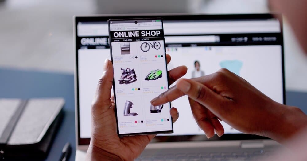 Man shopping on an e-commerce site on his phone with his computer in the background.