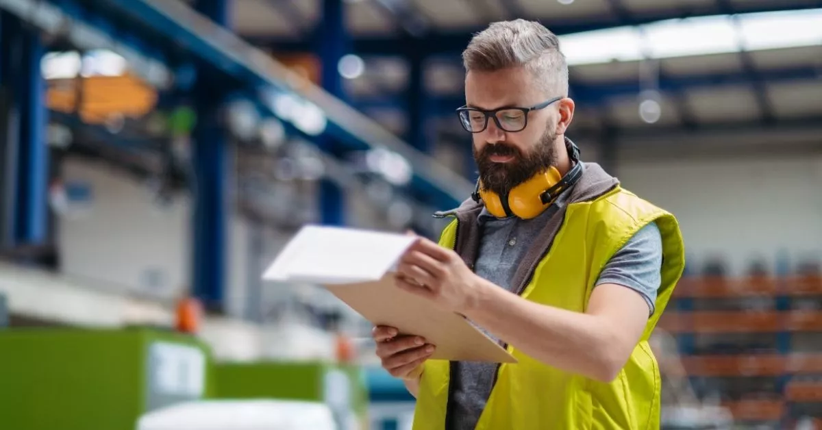 A warehouse worker wearing a safety vest and headphones reviews paperwork, checking details carefully as part of inventory or quality control