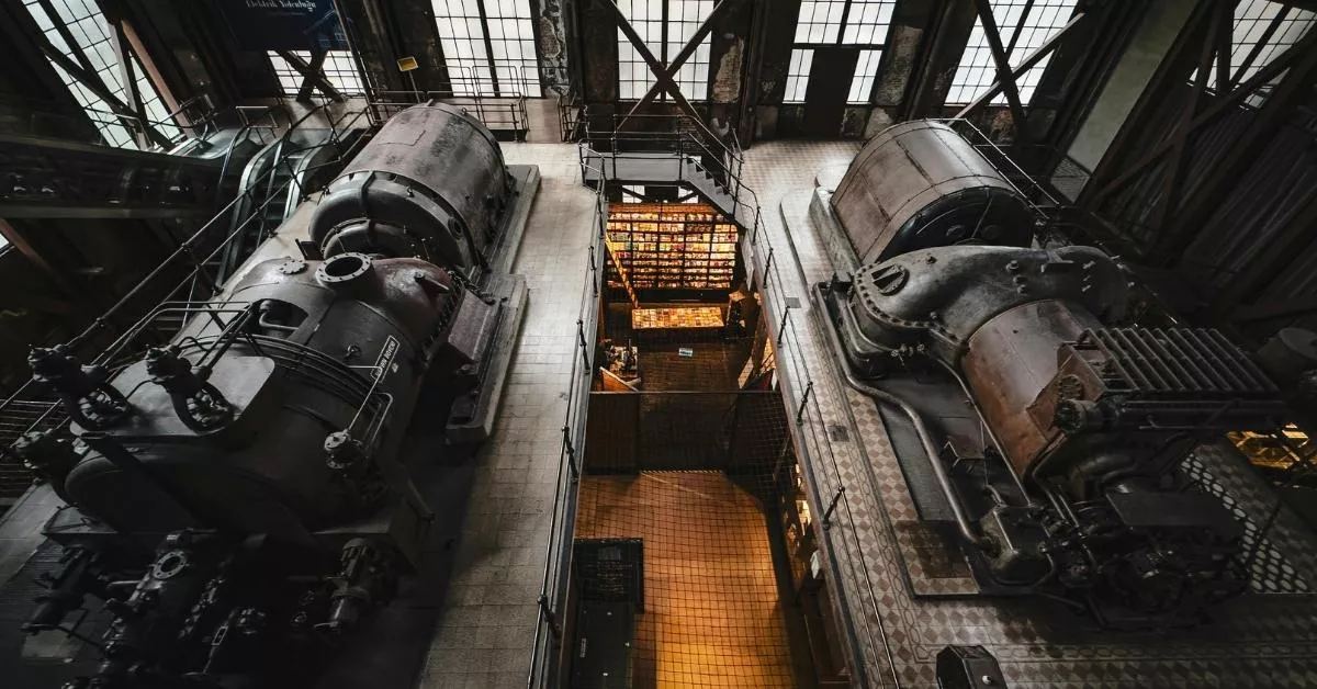 An overhead view of a large industrial facility with massive old machines and metal tanks lined up, giving off a gritty, heavy manufacturing vibe