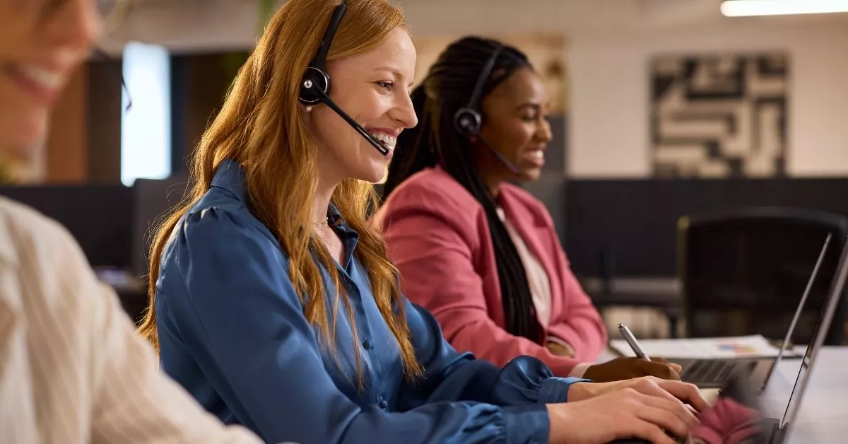 A group of customer support reps wearing headsets sit at computers, smiling and talking as they help people in a busy office