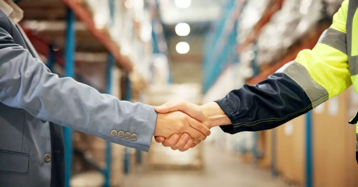 Two people shake hands in a warehouse aisle, showing a business deal or partnership coming together among shelves of packaged goods