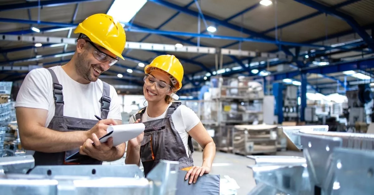 Two factory workers in safety gear stand together on a production floor, smiling while checking notes and reviewing work in a busy plant