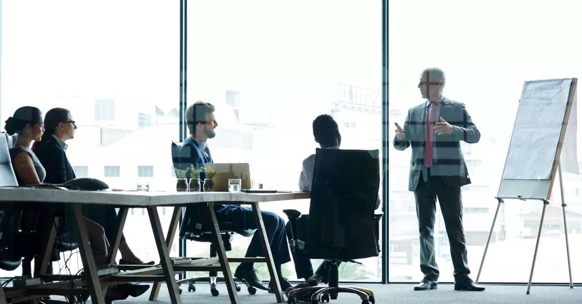Business team meeting with a presenter at a whiteboard, representing the collaborative product development process that includes design, engineering, sourcing, and manufacturing planning.