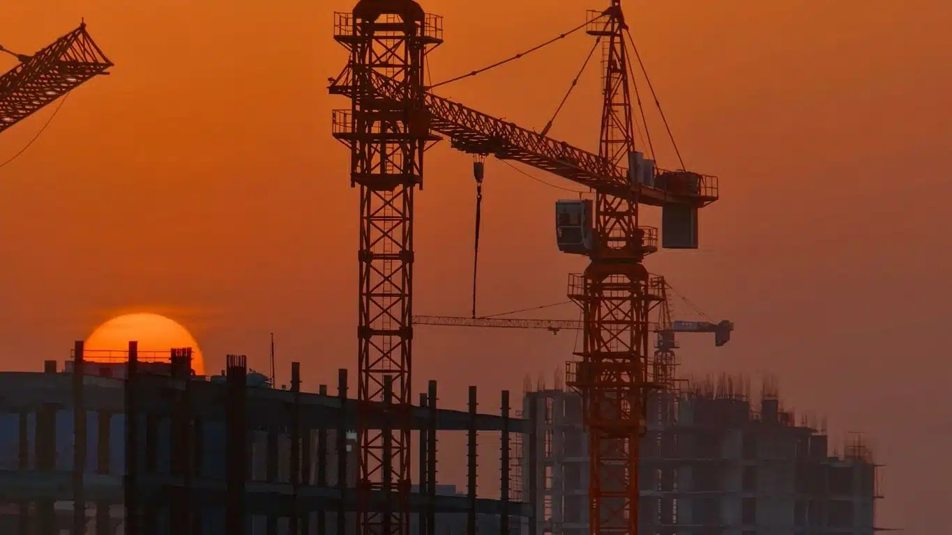 Tower cranes silhouetted over a construction site at sunset, the sun setting behind an unfinished concrete building